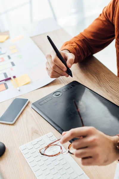 Enfoque selectivo del diseñador ux celebración de anteojos mientras se utiliza la tableta gráfica cerca de teléfono inteligente y diseños en la mesa - foto de stock