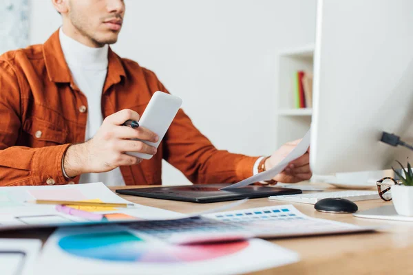 Zugeschnittene Ansicht des UX-Designers, der sein Smartphone hält, während er mit Webseiten-Skizzen und Farbpalette am Tisch im Büro arbeitet — Stockfoto