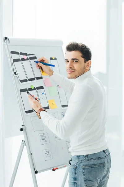 Side view of ux designer looking at camera while holding smartphone near mobile frameworks on whiteboard — Stock Photo