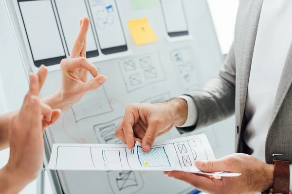 Cropped view of designer showing okay gesture near colleague with layouts of ux design near whiteboard in office — Stock Photo