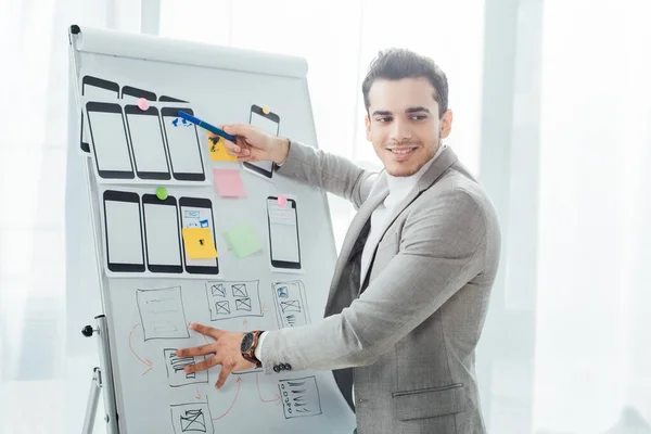 Sorrindo ux designer apontando com marcador em esboços do site e modelos móveis no quadro branco no escritório — Fotografia de Stock