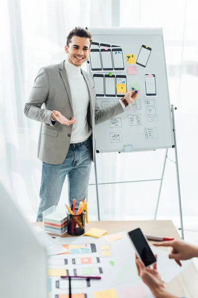 Enfoque selectivo del diseñador ux sonriente cerca de pizarra blanca con plantillas mirando a colega apuntando en el teléfono inteligente en la oficina - foto de stock