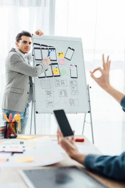 Selective focus of ux designer near whiteboard with layouts looking at colleague with smartphone showing okay sing in office — Stock Photo