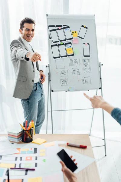 Foco seletivo de sorrir designer ux perto whiteboard com modelos apontando para colega com smartphone no escritório — Fotografia de Stock