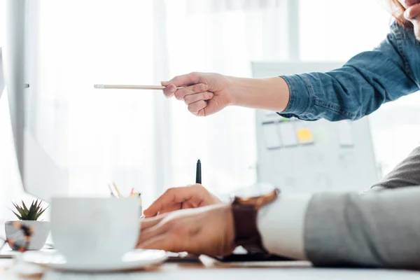 Selective focus of ux designer pointing on computer monitor to colleague at table in office — Stock Photo