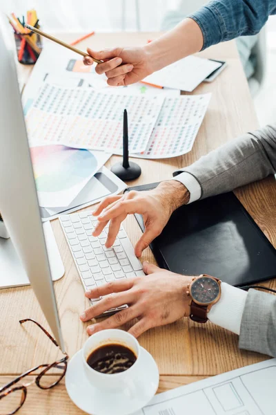 Vista cortada de designers usando o computador enquanto trabalhava no projeto de design de experiência do usuário perto de layouts e café na mesa — Fotografia de Stock