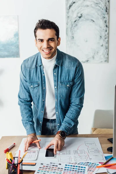 Lächelnder Designer blickt in die Kamera, während er mit UX-Wireframe-Skizzen, Farbpaletten und Smartphone auf dem Tisch arbeitet — Stockfoto