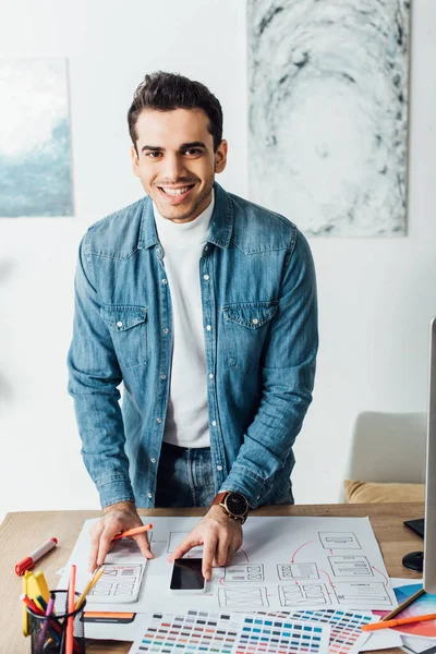 Designer souriant regardant la caméra tout en travaillant avec des croquis ux wireframe, palettes de couleurs et smartphone sur la table — Photo de stock