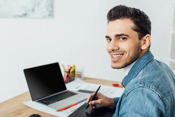Smiling ux diseñador mirando a la cámara mientras se utiliza la tableta gráfica y el ordenador portátil en la mesa - foto de stock