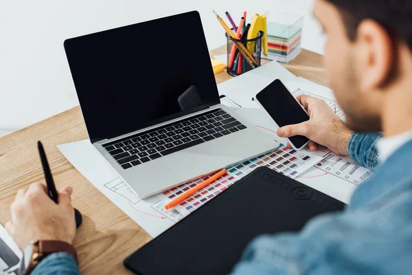 Foco seletivo do designer ux usando smartphone perto de laptop, tablet gráficos e layouts com esboços na mesa em fundo cinza — Fotografia de Stock