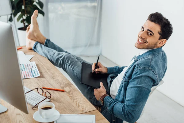Barfuß-UX-Designer lächelt in die Kamera, während er Grafik-Tablet und Computer in der Nähe der Farbpalette auf dem Tisch im Büro benutzt — Stockfoto