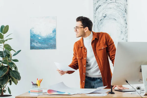 Diseñador sonriente mirando hacia otro lado mientras trabaja en el proyecto de diseño ux cerca de círculos de color y bocetos en la mesa - foto de stock