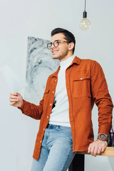 Vista de ángulo bajo del papel de diseñador ux sonriente y mirando hacia otro lado cerca de la mesa de trabajo - foto de stock