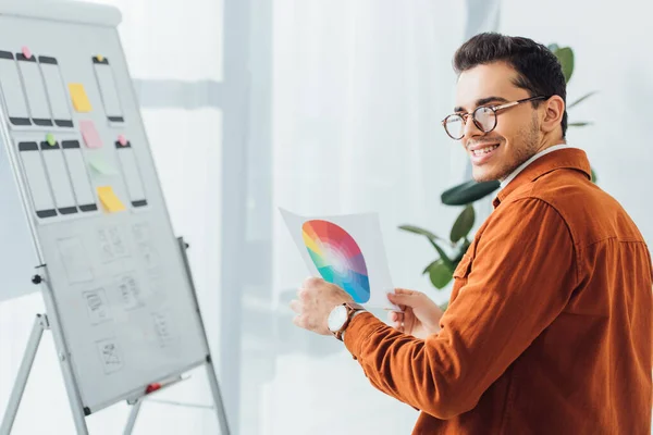 Selective focus of smiling ux designer holding color circle near layouts of mobile website design on whiteboard in office — Stock Photo