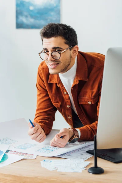 Enfoque selectivo del diseñador ux sonriendo a la cámara cerca del monitor de la computadora y bocetos del wireframe del sitio web en la mesa - foto de stock