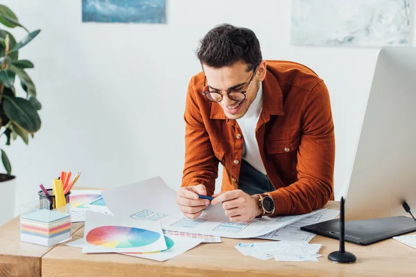 Cheerful ux designer working with website wireframe sketches and layouts near computer monitor and graphics tablet on table — Stock Photo