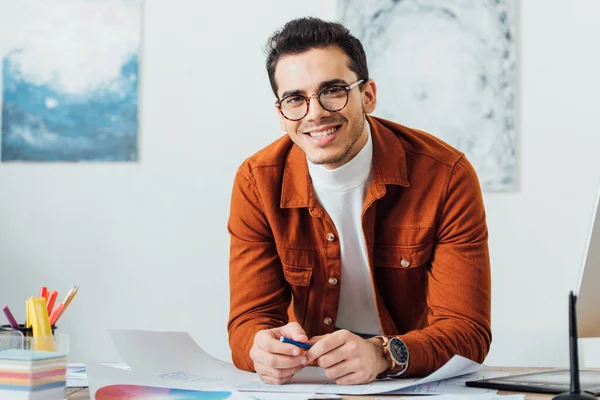 Designer créatif souriant à la caméra tout en travaillant sur le projet de conception de l'expérience utilisateur à la table dans le bureau — Photo de stock