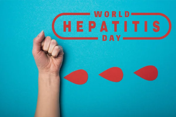 Vue du dessus des gouttes de papier de sang près du poing féminin sur fond bleu, illustration de la journée mondiale de l'hépatite — Photo de stock