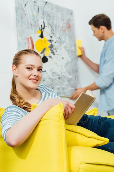 Foco seletivo da menina com livro sorrindo para a câmera perto namorado limpeza pintura na sala de estar — Fotografia de Stock