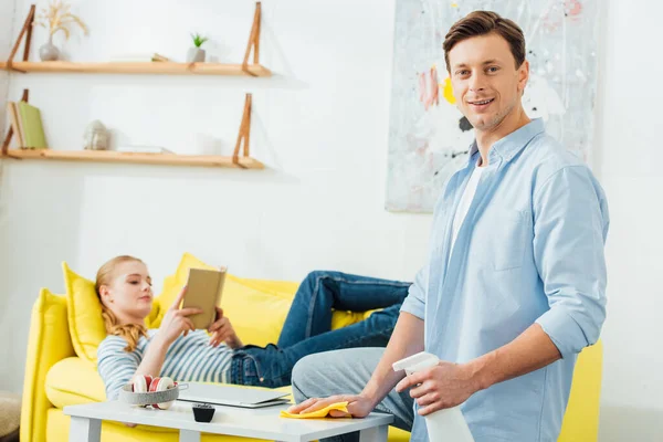 Selective focus of smiling man cleaning coffee table with detergent and rag near girlfriend reading book on couch in living room — Stock Photo