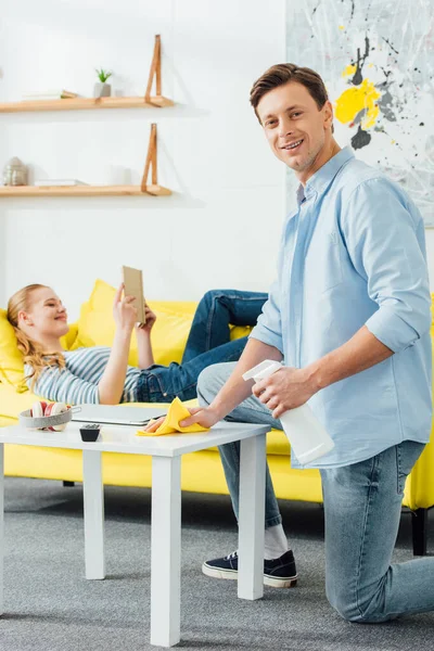Vista lateral do homem sorridente com pano e detergente limpeza mesa de café perto da namorada com livro no sofá em casa — Fotografia de Stock