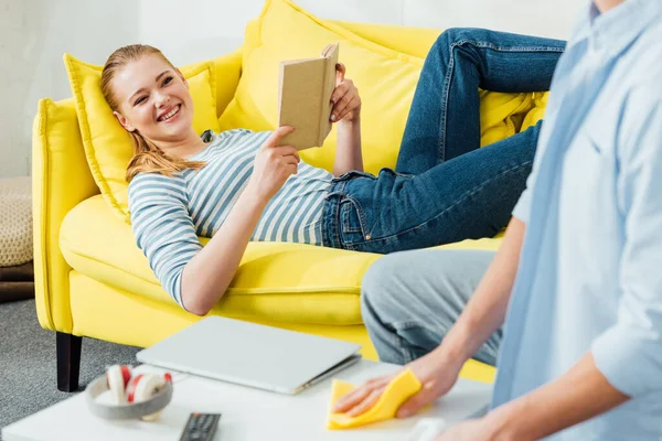 Selektiver Fokus einer lächelnden Frau mit Buch, die ihren Freund beim Putzen des Wohnzimmertisches beobachtet — Stockfoto