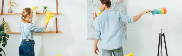 Back view of young couple cleaning with dust brush and spray detergent in living room, panoramic shot — Stock Photo