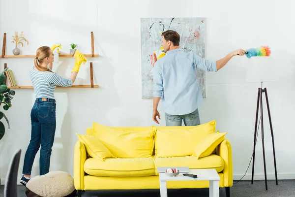 Back view of woman cleaning shelf near boyfriend with dust brush in living room — Stock Photo