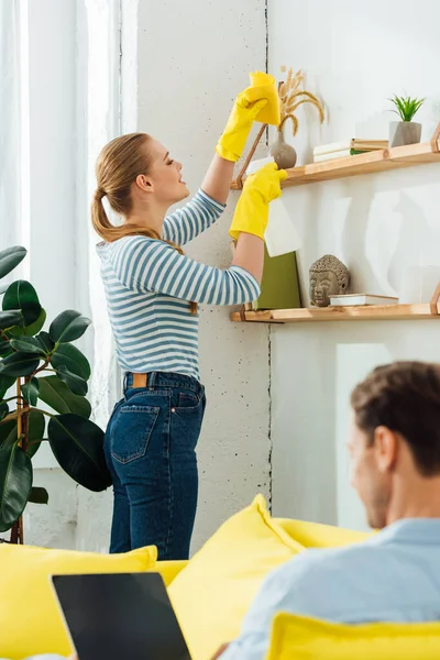 Selective focus of girl with rag and bottle of detergent cleaning shelf near boyfriend with laptop on couch — Stock Photo