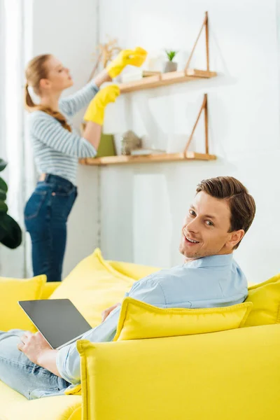 Selektiver Fokus eines Mannes, der in die Kamera lächelt, während er Laptop in der Nähe der Freundin benutzt, die Regal im Wohnzimmer putzt — Stockfoto