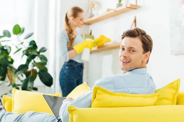 Enfoque selectivo del hombre con el ordenador portátil sonriendo a la cámara mientras está sentado en el sofá cerca de la novia estante de limpieza en la sala de estar - foto de stock