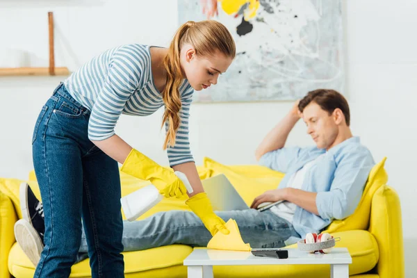 Vista laterale della ragazza che pulisce il tavolino con detersivo e straccio vicino al fidanzato usando il computer portatile sul divano in soggiorno — Foto stock