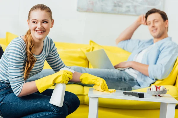 Selektiver Fokus der Frau, die weglächelt, während sie Couchtisch mit Waschmittel putzt und Lappen in der Nähe ihres Freundes mit Laptop auf der Couch — Stockfoto