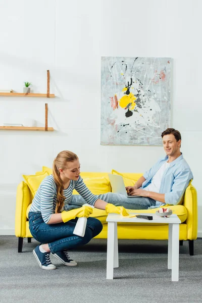 Vue latérale de l'homme souriant avec ordinateur portable regardant petite amie nettoyage table basse dans le salon — Photo de stock