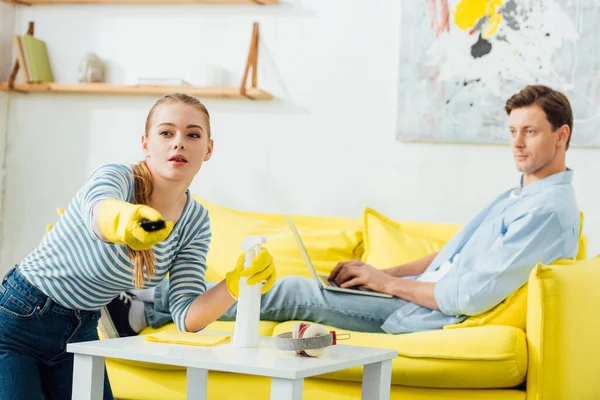 Selective focus of woman with cleaning supplies using remote controller near boyfriend with laptop on couch at home — Stock Photo