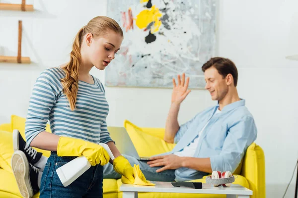 Selective focus of girl cleaning coffee table with rag and detergent near boyfriend having video chat on laptop on couch at home — Stock Photo
