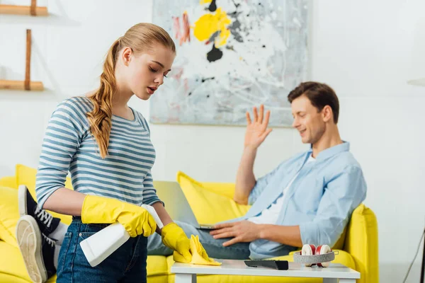 Focus selettivo di ragazza pulizia tavolino con straccio e detergente vicino fidanzato avere video chat sul computer portatile sul divano a casa — Foto stock