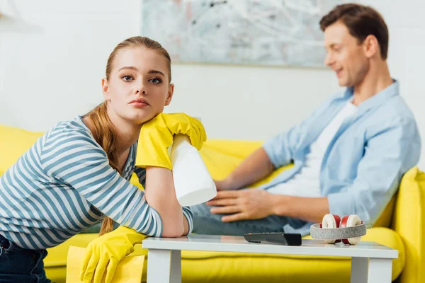 Concentration sélective de la femme avec détergent et chiffon regardant la caméra près du petit ami en utilisant un ordinateur portable sur le canapé dans le salon — Photo de stock