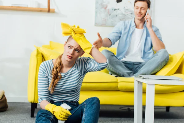 Enfoque selectivo de la mujer cansada con suministros de limpieza sentado en el suelo cerca de novio hablando en el teléfono inteligente en el sofá en casa - foto de stock