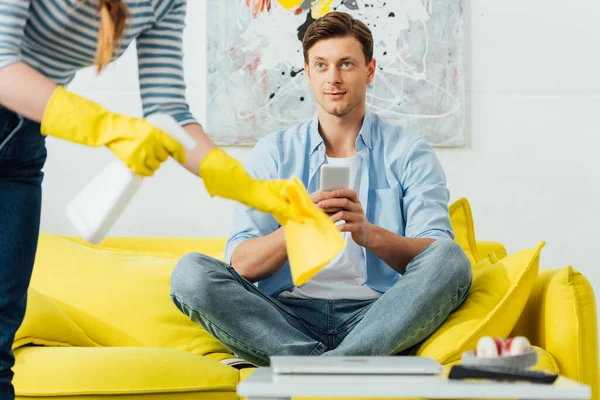 Selektiver Fokus des Mannes mit Smartphone auf Freundin, die Couchtisch im Wohnzimmer putzt — Stockfoto