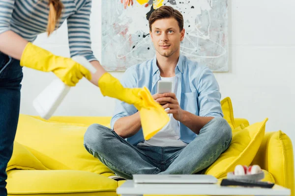 Selective focus of man with smartphone looking at girlfriend cleaning coffee table in living room — Stock Photo