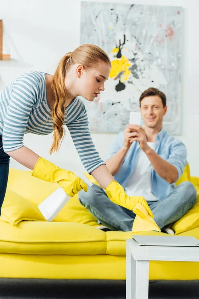 Enfoque selectivo de la mujer con detergente y trapo de limpieza de mesa de café cerca de novio con smartphone en el sofá en la sala de estar - foto de stock