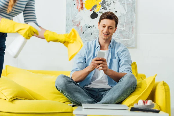 Focus selettivo di uomo sorridente utilizzando smartphone sul divano vicino alla donna con detersivo e straccio in soggiorno — Foto stock