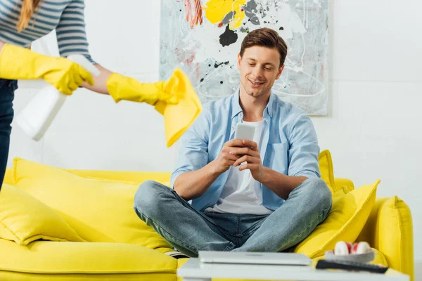 Selective focus of smiling man using smartphone on couch near woman with detergent and rag in living room — Stock Photo