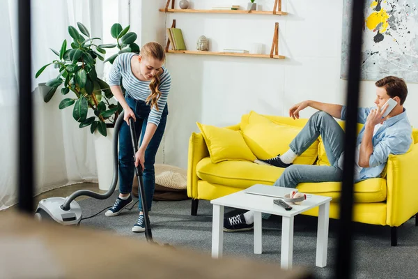 Focus sélectif de l'homme parlant sur smartphone tandis que la petite amie nettoyage tapis avec aspirateur dans le salon — Photo de stock