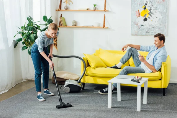 Hombre usando teléfono inteligente mientras su novia limpieza de alfombras con aspiradora en la sala de estar - foto de stock