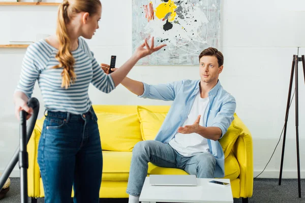 Selective focus of woman with vacuum cleaner quarreling with boyfriend holding remote controller on couch — Stock Photo