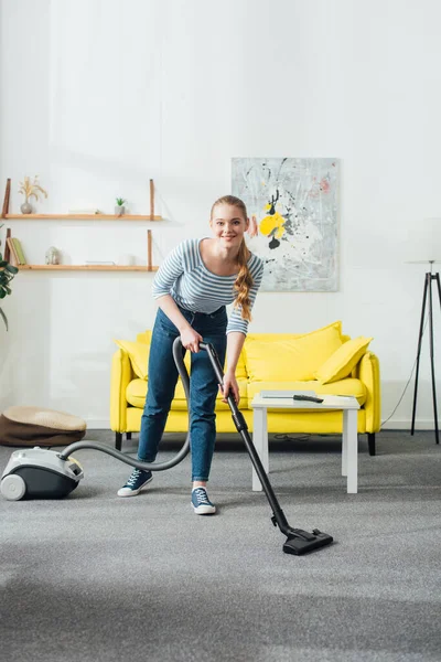 Mujer atractiva sonriendo a la cámara mientras limpia la alfombra con aspiradora en la sala de estar - foto de stock