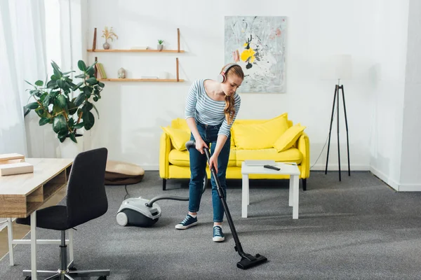 Jeune femme dans les écouteurs nettoyage tapis avec aspirateur dans le salon — Photo de stock