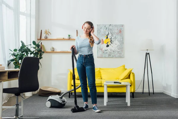 Attraktiv lächelnde Frau mit Kopfhörer putzt Teppich zu Hause mit Staubsauger — Stockfoto